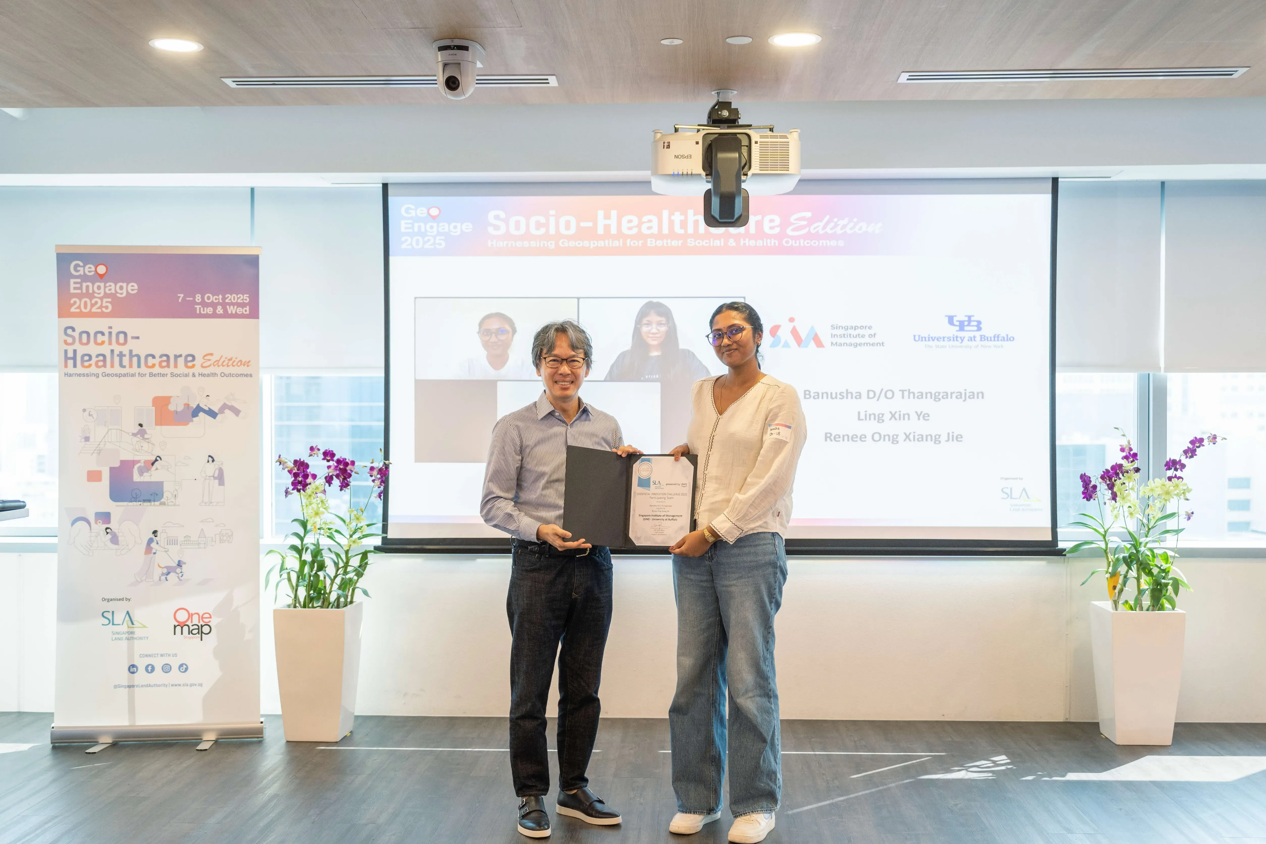 Certificate presentation for SIM for AWS-SLA Geospatial Innovation Challenge at GeoEngage 2025 Socio-Healthcare Edition event, with a presentation screen behind them.